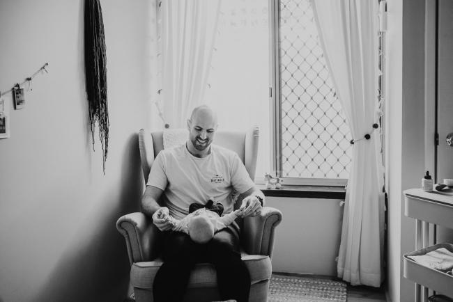 Black and white image of father holding baby on chair in nursery with Perth Newborn photographer Alana Prosper Photography