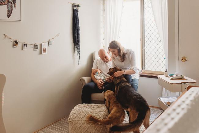 Parents on chair in nursery with baby, and dogs in the room with Perth Newborn photographer Alana Prosper Photography