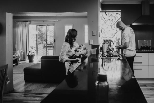 Black and white image of mother holding baby in kitchen while father gets bottle ready with Perth Newborn photographer Alana Prosper Photography