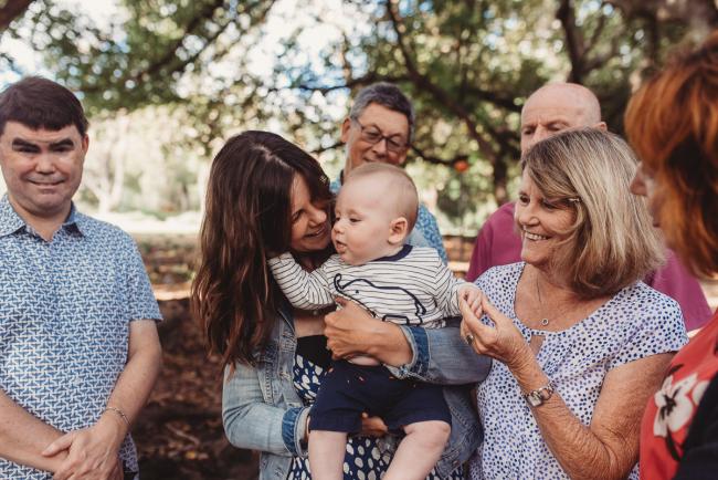 Mother holding baby surrounded by family during extended family session at Hyde Park with Perth family photographer Alana Prosper Photography