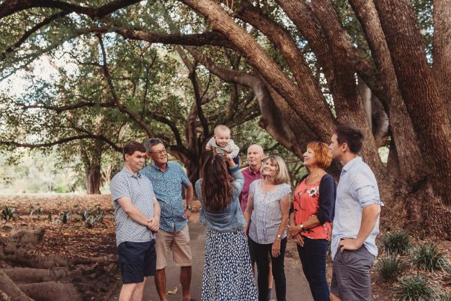 Mother lifting baby up in the air surrounded by family during extended family session at Hyde Park with Perth family photographer Alana Prosper Photography