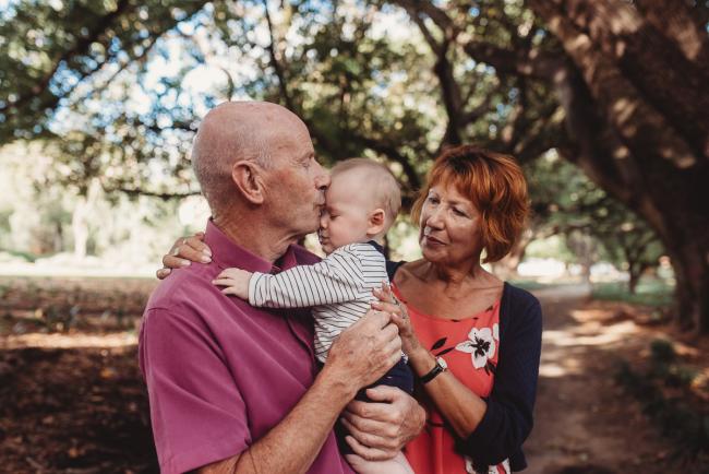 Grandfather holding and kissing baby as grandmother cuddles them during extended family session at Hyde Park with Perth family photographer Alana Prosper Photography