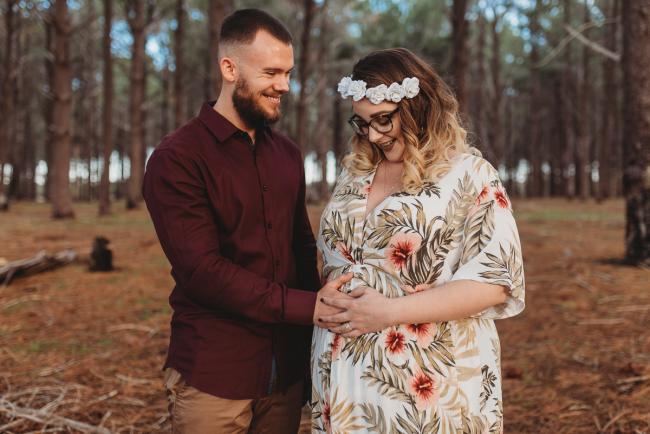 Couple smiling and looking down at her tummy at The Pines with Perth maternity photographer Alana Prosper Photography