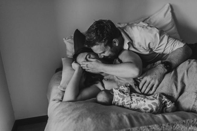 black and white image of mother laughing and feeding baby on bed as partner kisses her with Perth newborn lifestyle photographer Alana Prosper Photography