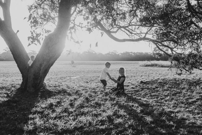 black and white image of mother kneeling on the ground holding sons hand with other son in a tree during golden hour at Perry's Paddock with Perth Family Photographer Alana Prosper Photography