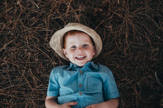 Little boy in hat laying on the ground and smiling during Perth family photography session at The Pines Perth with Alana Prosper Photography