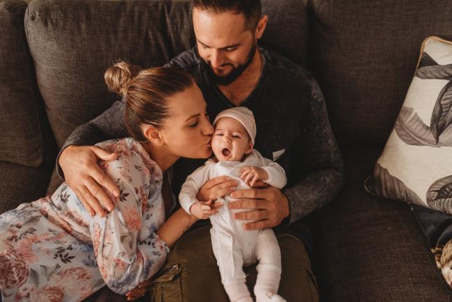 Father holding newborn baby on lap as mother kisses baby with Perth newborn lifestyle photographer