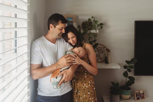 mother and father stand by window with baby with Perth Newborn photographer