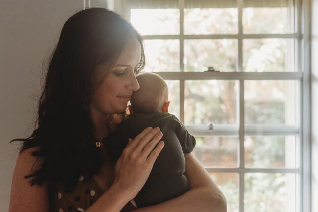 mother holding baby on shoulder by window with Perth Newborn photographer